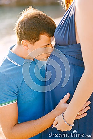 Man kissing pregnant belly of woman in long blue dress on sunny day Stock Photo