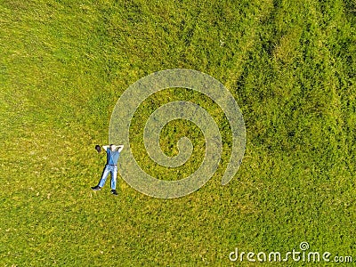 Man lying on the ground in a park on a green grass field. Man dressed in blue t shirt and jeans hands behind hes head. Aerial top Stock Photo