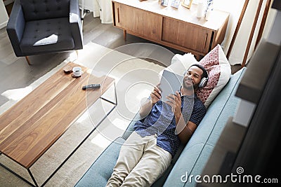 Man Lying On Sofa At Home Wearing Headphones And Watching Movie On Digital Tablet Stock Photo