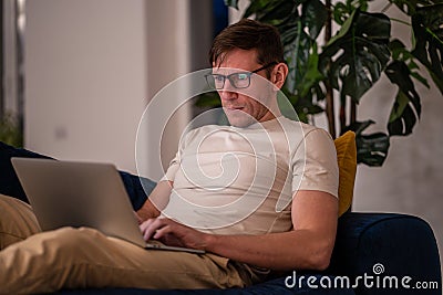 Man middle aged student working on educational project lying on couch at home in evening after job. Stock Photo