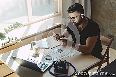 Man millenial in glasses sits at table in front of laptop and uses smartphone, reads message from business partner Stock Photo