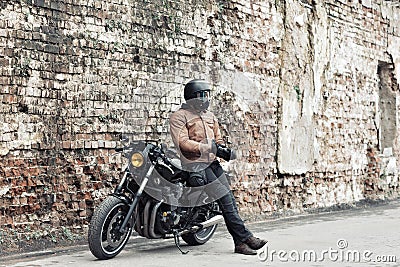 Man a motorcyclist standing with helmet near her bike, brick wall of garage background. Male in a leather jacket and Stock Photo