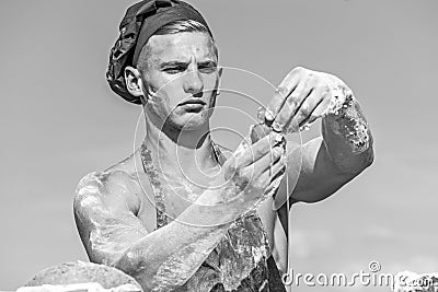 Man muscular baker or cook covered with flour working outdoor, sky on background. Baker adds egg to prepare dough Stock Photo