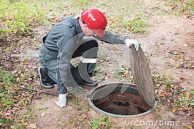 a man in overalls opened a sewer hatch and looks into a septic tank. Cleaning of sewers and drains Stock Photo