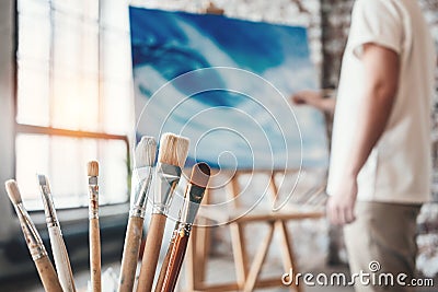 Man painter working in workshop with canvas on easel. Brushes close up on wooden table in studio. Flare effect Stock Photo