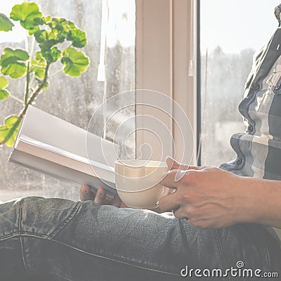 A man in a plaid shirt and jeans sits on a window sill with a cup of coffee and a notepad Stock Photo