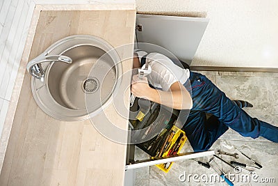 Man plumber in uniform fixing the sink with adjustable spanner in his hand lying on the kitchen floor professional Stock Photo