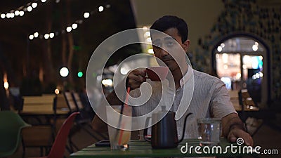 Man pours boiling water into mug at cafe. Media. Attractive man drinks coffee at table near cafe with evening lights Stock Photo