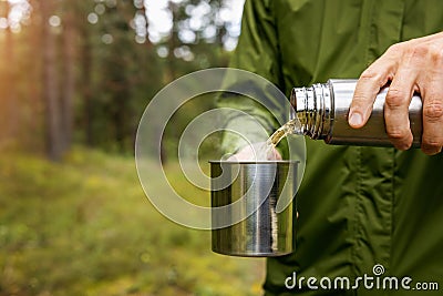 man pours hot drink from thermos flask into a metal cup in forest. nature tourism and camping gear concept Stock Photo