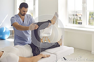 Man professional chiropractor in blue uniform fixing and setting lying man patients legs joints Stock Photo