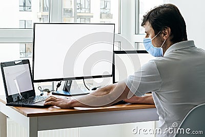 A man in protective blue medical mask is sitting at a workplace with two laptops and a monitor near the window. Office work Stock Photo