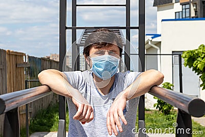 Man in protective blue medical mask standing on the outdoor sports ground, resting his hands on the bars. Male in a respirator Stock Photo