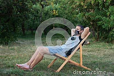 Man in protective medical mask on his face sunbathing outside green lawn in park. Person outdoors relaxing on deck chair in garden Stock Photo