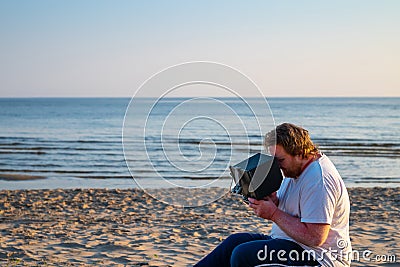 Man With Psoriasis On The Skin Of His Hands Sits On The Beach At Sunrise And Controls The Drone Through The Remote Control With Stock Photo