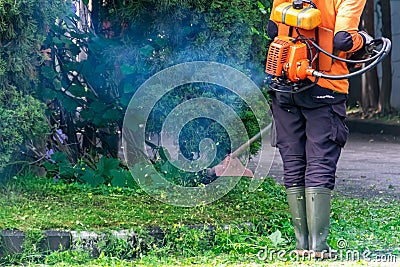 Man public worker tidy up the city street yard with the grass cutter or trimmer machine with a smoke blow away Stock Photo