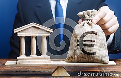 A man puts a euro money bag on scales opposite to government building, bank, university. Taxes payment. Lobbying interests. Stock Photo