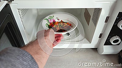 A man puts his hand a white bowl with a pattern with food in the microwave front view, open microwave Stock Photo