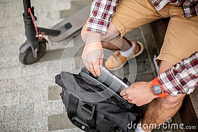 Man Putting Laptop in Backpack outside sitting near electric transport scooter. Caucasian man packing laptop in backpack. Male Stock Photo