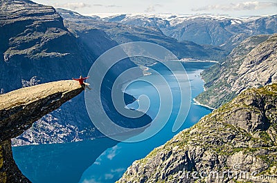 A man in a red jacket sitting on the Trolltunga rock with a blue lake 700 meters lower and interesting sky with clouds Stock Photo