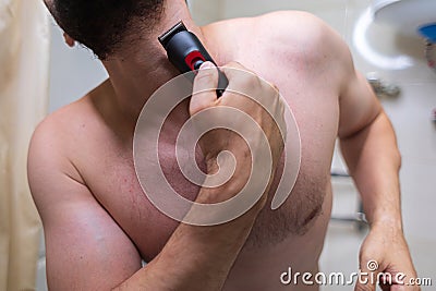 A man removes hair from his beard with an electric shaver. A man with an athletic figure in the bathroom Stock Photo