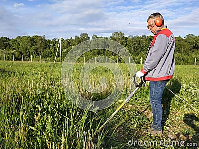 A man rows grass with an oak trimmer in the field, in the yard is summer and the sun shines Stock Photo