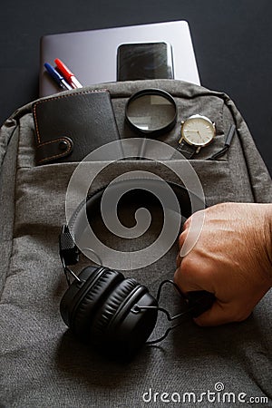 A man`s hand holds wireless headphones on the background of a gray textile backpack with accessories and gadgets for a trip or Stock Photo