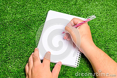 The man`s hand was holding a pen to write something down in a notebook which is on the green grass Stock Photo