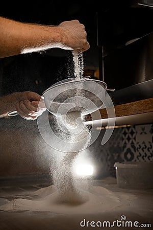 Man's hands sift flour through a metal sieve in a dark kitchen Stock Photo