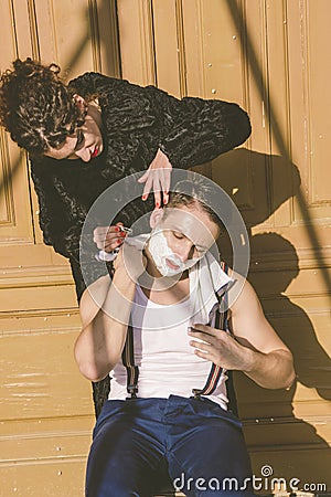 man with shaving foam on his face and towel around his neck siting on chair and posing while pretty women shave him Stock Photo