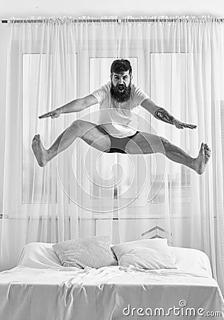Man in shirt and underpants jumping on bed, white curtains on background. Macho with beard jumps high in air. Guy on Stock Photo
