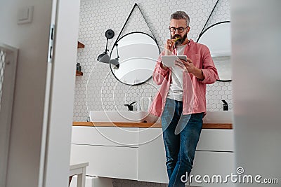 Man shopping online while brushing his teeth in bathroom. Holding tablet, scrolling and buying in eshop, toothbrush in Stock Photo