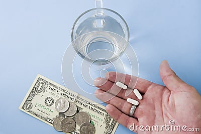 Man showing hand with white pills above light pastel blue background desk with money, coins and a glass of fresh water. Stock Photo