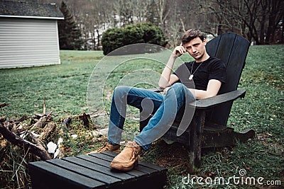 Man sits outdoors on a wooden chair on the backyard of his house at rural area. Stock Photo