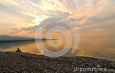 A man sitting alone on a log at the over the edge of a sea, dawn silence on lake. Editorial Stock Photo