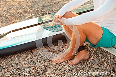 A man sitting at the pebble beach with a sup board. Feet and surfboard close up. Summer recreation and activity Stock Photo