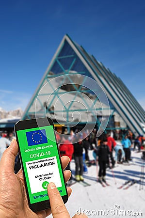 A man on a ski slope near the cable car station and ski lifts holds a smartphone with the European Union digital green pass for Stock Photo