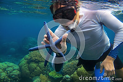 Man snorkeling in blue water with star fish. Snorkeling in coral reef. Snorkel holds blue starfish. Stock Photo