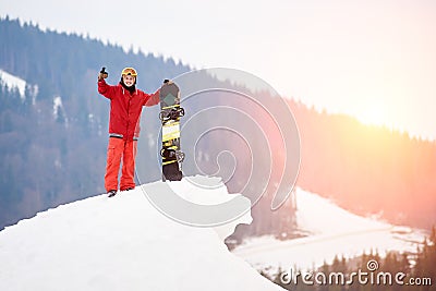 Man snowboarder standing on the top of the snowy slope with snowboard, showing thumbs up at winter ski resort Stock Photo