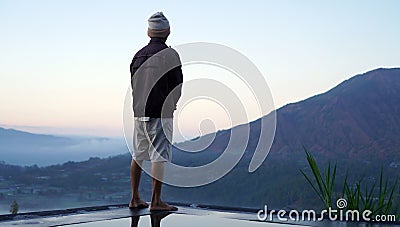 Man standing alone from behind on the edge of the pool at sunrise with water reflection and blue mount Batur view background. Stock Photo