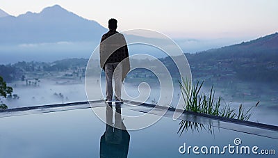 Man standing alone from behind on the edge of the pool at sunrise with water reflection and blue mount Batur view background. Stock Photo