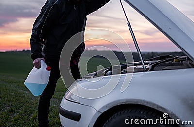 Man standing near car with open hood and holding a bottle with blue fluid on sunset. Car breakdown. Confused man stands Stock Photo