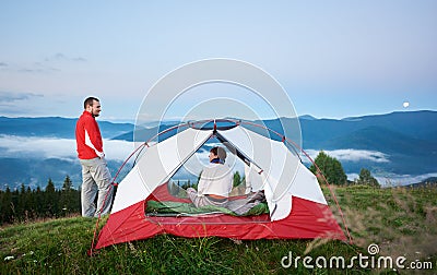 Man standing near the tent in which sits woman against beautiful scenery of mighty mountains Stock Photo