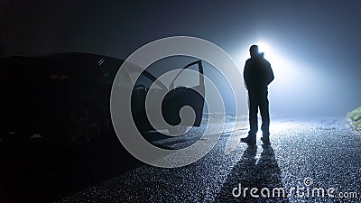 A man standing next to a car, with door open, parked on the side of the road, underneath a street light, on a spooky, scary, rural Stock Photo