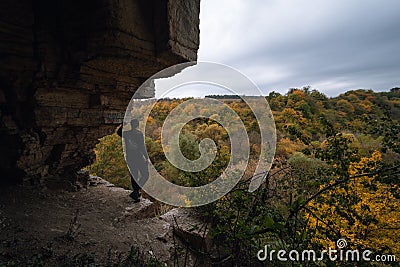 A man stands on the edge, at the exit from the cave. View of the autumn forest, tree crowns. Dramatic gray clouds. Travel and Stock Photo