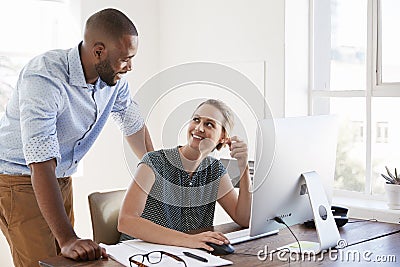 Man stands talking to woman smiling at her desk in an office Stock Photo