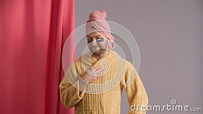 A man strokes his face and beard in the studio after a shower against a pink shower curtain. A man in yellow robe, pink Stock Photo