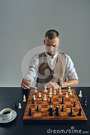 Man with stylish mustache, dressed in brown vest, white shirt is playing chess. Nearby is a cup of coffee. Grey Stock Photo