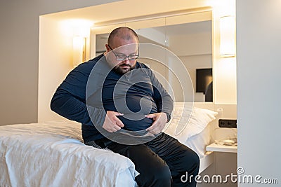 Man suffering from extra weight touching abdomen sitting on bed in bedroom. Stock Photo