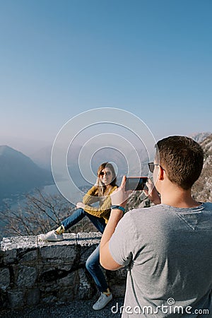 Man takes a picture with a smartphone of woman sitting on a fence over the Bay of Kotor. Montenegro. Back view Stock Photo