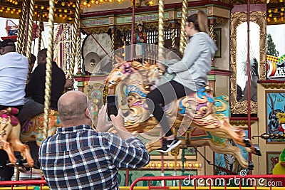 Man taking a photo on a mobile phone of teenager riding on a carousel horse on fairground ride at amusement fair Editorial Stock Photo